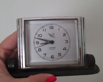 May include: A silver alarm clock with a black base. The clock face is white with black numbers and hands. The clock says "Pioneer Сделано в СССР".