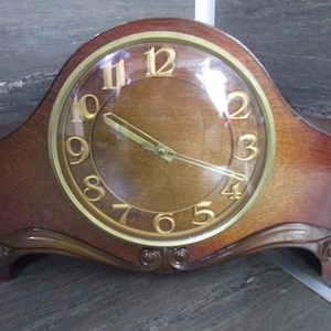 May include: A vintage wooden mantel clock with a gold-toned face and black hands. The clock has a curved, ornate base and a glass cover over the face.