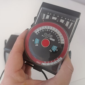 May include: A black and red light meter with a circular dial labeled "cds WEIMAR" and a smaller dial labeled "ASA" and "DIN".