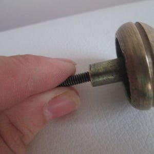 Puede incluir: Un pomo de armario plateado con un tornillo adjunto. El pomo tiene una forma redondeada y abovedada con un anillo decorativo elevado alrededor del borde.