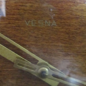 May include: Close-up of a wooden clock face with a gold-coloured hand and the word "VESNA" inscribed on the surface.