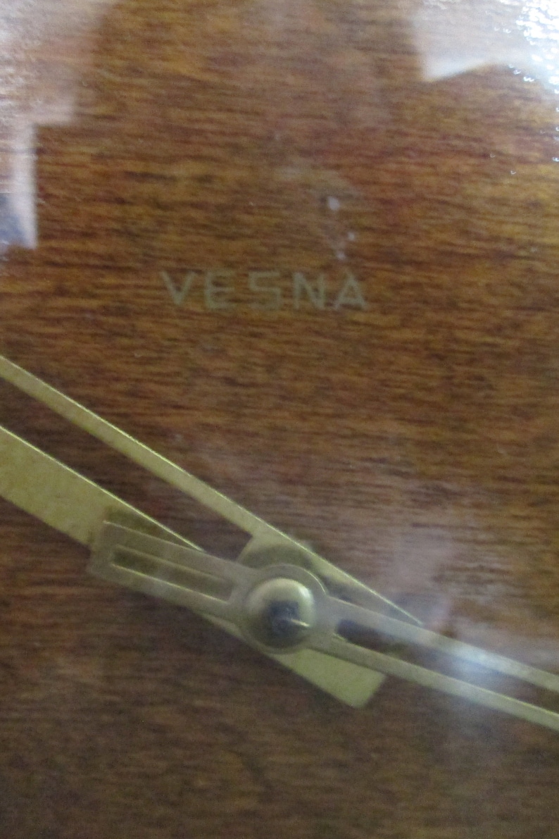 May include: Close-up of a wooden clock face with a gold-coloured hand and the word "VESNA" inscribed on the surface.