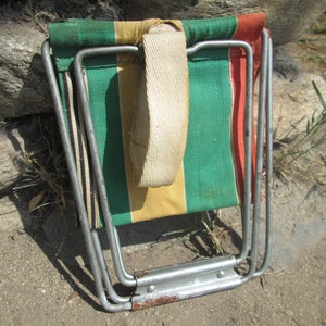 K&ouml;nnte beinhalten: Ein zusammengeklappter, gestreifter Campingstuhl aus Segeltuch mit einem wei&szlig;en Trageriemen und einem silbernen Metallrahmen.