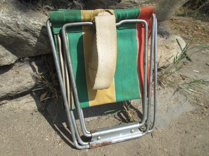 K&ouml;nnte beinhalten: Ein zusammengeklappter, gestreifter Campingstuhl aus Segeltuch mit einem wei&szlig;en Trageriemen und einem silbernen Metallrahmen.
