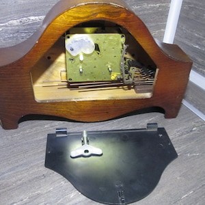 May include: The back of a wooden clock with the clockwork mechanism exposed. The clock face is missing. The back of the clock is made of dark wood and has a curved shape. The clockwork mechanism is made of brass and has a number of gears and springs. The clock is missing its face and hands.
