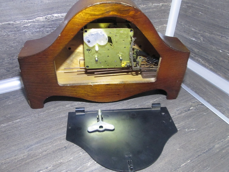 May include: The back of a wooden clock with the clockwork mechanism exposed. The clock face is missing. The back of the clock is made of dark wood and has a curved shape. The clockwork mechanism is made of brass and has a number of gears and springs. The clock is missing its face and hands.