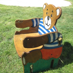 May include: A wooden children's chair shaped like a bear wearing a blue and white striped shirt and orange shorts. The chair is painted brown, blue, white, orange, and green.