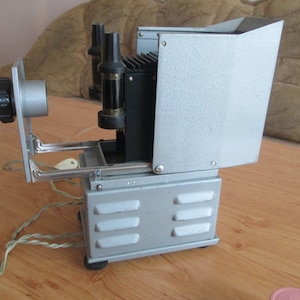 Puede incluir: Un proyector de diapositivas vintage plateado con una lente negra y dos bombillas visibles. El proyector está sobre una superficie de madera.