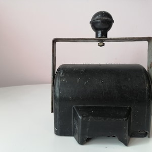 Op de afbeelding: Een vintage zwarte stempel met een metalen handvat en een afgeronde bovenkant. De stempel heeft een rechthoekige basis en een cilindrisch lichaam. Het handvat is bevestigd aan de bovenkant van de stempel. De stempel staat op een wit oppervlak.
