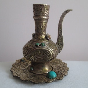 May include: An ornate silver-coloured metal pitcher with a spout and a decorative base. The pitcher has intricate carvings and is adorned with turquoise-coloured stones.