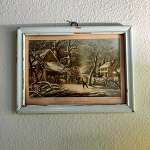 Puede incluir: Una impresión de arte mural rectangular titulada A SNOWY MORNING, que muestra un paisaje invernal con un granero, árboles y figuras, encerrada en un marco de madera azul claro desgastado con desconchones de pintura y un gancho metálico.