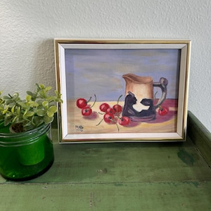 May include: Framed still life painting featuring a tan and black pitcher with cherries. The painting has a gold frame and is signed "M.C." in the lower left corner. The painting is on a green surface.