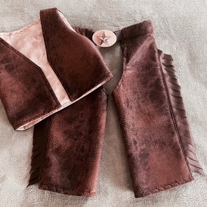 May include: A brown faux leather vest and matching chaps set with a textured finish. The vest features a light-colored interior lining. The chaps include side fringe detailing and a central belt buckle with a prominent star emblem in the middle.