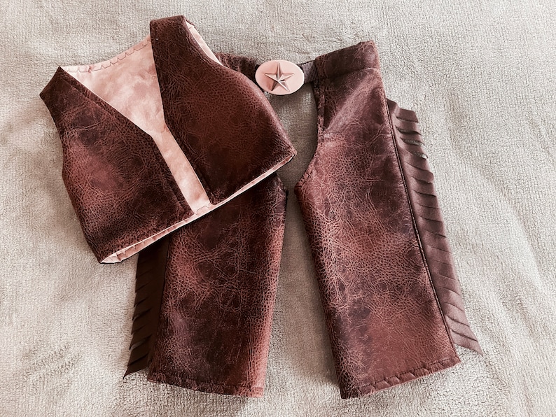 May include: A brown faux leather vest and matching chaps set with a textured finish. The vest features a light-colored interior lining. The chaps include side fringe detailing and a central belt buckle with a prominent star emblem in the middle.
