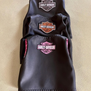 Puede incluir: Tres fundas de asiento de motocicleta negras apiladas una encima de la otra. Cada funda tiene un parche con el logotipo de Harley-Davidson en el centro. El parche superior es blanco y negro, el del medio es naranja y negro, y el inferior es rosa y negro.