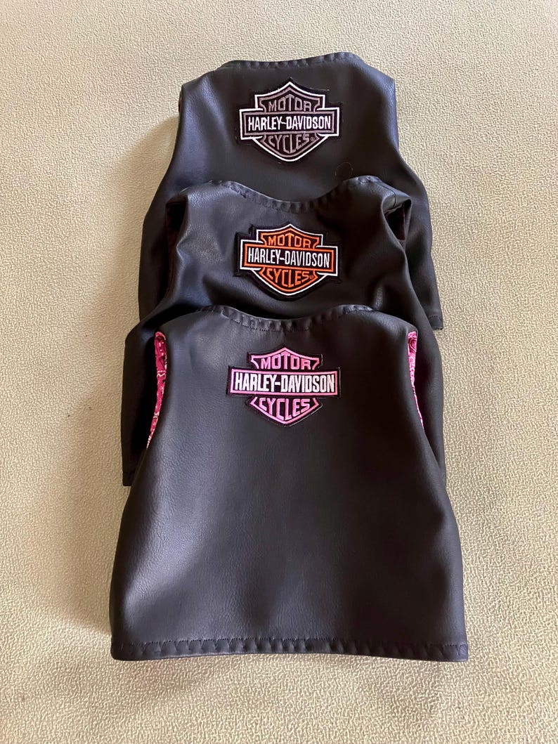 Puede incluir: Tres fundas de asiento de motocicleta negras apiladas una encima de la otra. Cada funda tiene un parche con el logotipo de Harley-Davidson en el centro. El parche superior es blanco y negro, el del medio es naranja y negro, y el inferior es rosa y negro.