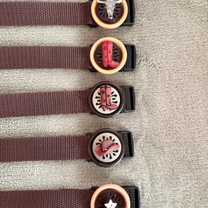 May include: Five brown nylon webbing straps with circular plastic buckles. Each buckle features a unique decorative center piece: a silver steer skull, a red boot, a pink boot, a brown hat, and a white star, all set against a dark cutout background.