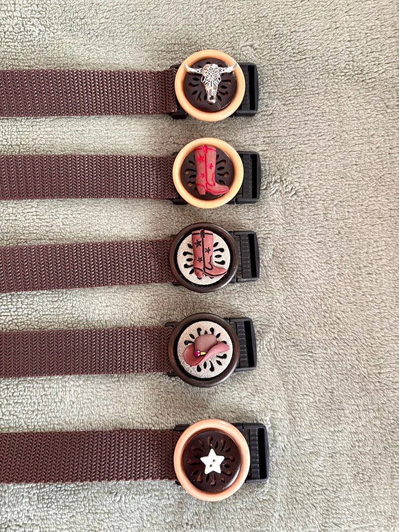 May include: Five brown nylon webbing straps with circular plastic buckles. Each buckle features a unique decorative center piece: a silver steer skull, a red boot, a pink boot, a brown hat, and a white star, all set against a dark cutout background.