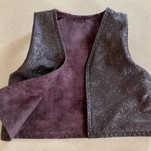 Puede incluir: Un chaleco marr&oacute;n oscuro con un estampado floral en relieve en el exterior. El interior es de un color p&uacute;rpura apagado. El chaleco tiene cuello en V y est&aacute; abierto por delante.