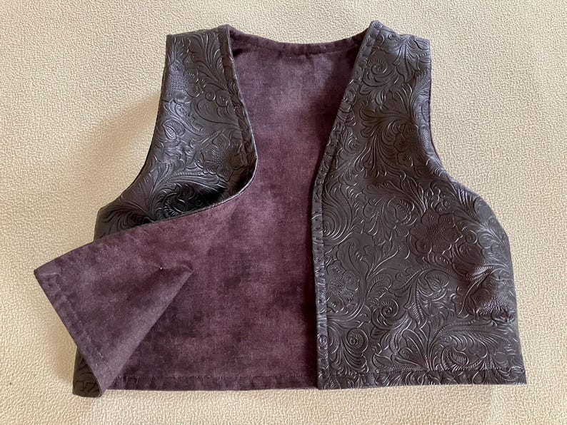 Puede incluir: Un chaleco marr&oacute;n oscuro con un estampado floral en relieve en el exterior. El interior es de un color p&uacute;rpura apagado. El chaleco tiene cuello en V y est&aacute; abierto por delante.