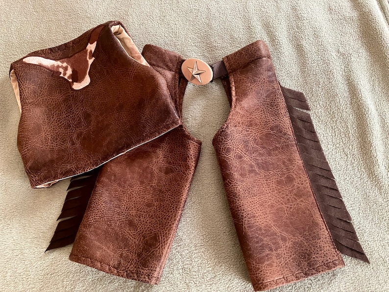 May include: A set of brown faux leather chaps and a matching vest featuring a textured finish. The chaps include dark brown fringe along the outer edges and a central belt buckle with a silver star emblem. The vest displays a cowhide pattern.