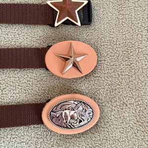 May include: Three brown woven belts with distinct buckles. The top buckle is a brown star with a gold border. The middle buckle is an oval light tan leather piece with a gold star. The bottom buckle is an oval light tan leather piece with a silver horse.