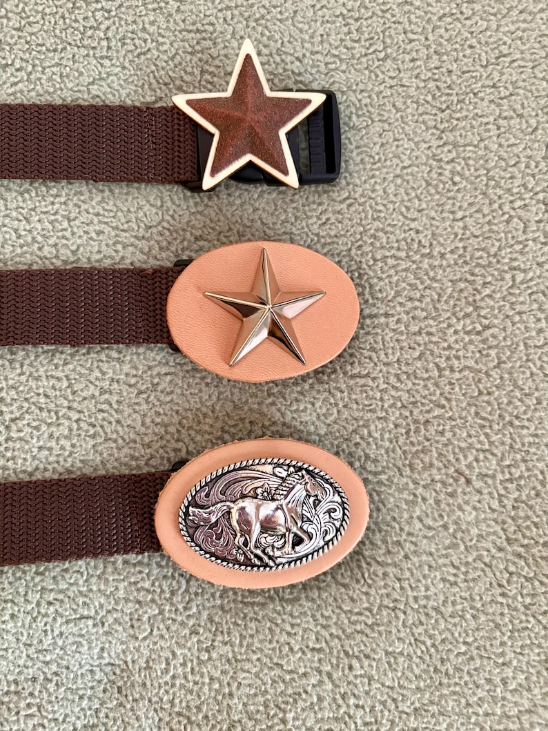 May include: Three brown woven belts with distinct buckles. The top buckle is a brown star with a gold border. The middle buckle is an oval light tan leather piece with a gold star. The bottom buckle is an oval light tan leather piece with a silver horse.