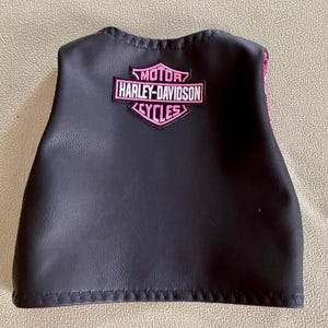 Puede incluir: Chaleco de cuero negro con un parche rosa con el logotipo de Harley-Davidson Cycles. El parche presenta el nombre de la marca en una fuente estilizada. El chaleco tiene una forma curva y es probablemente para un ni&ntilde;o o un adulto peque&ntilde;o.