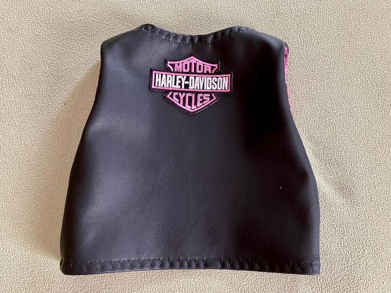 Puede incluir: Chaleco de cuero negro con un parche rosa con el logotipo de Harley-Davidson Cycles. El parche presenta el nombre de la marca en una fuente estilizada. El chaleco tiene una forma curva y es probablemente para un ni&ntilde;o o un adulto peque&ntilde;o.