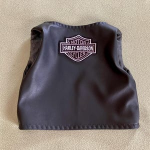 Puede incluir: Un chaleco de cuero negro con el parche del logotipo de Harley-Davidson Cycles. El parche es blanco, negro y rosa. El chaleco tiene un dobladillo cosido y un forro interior suave.