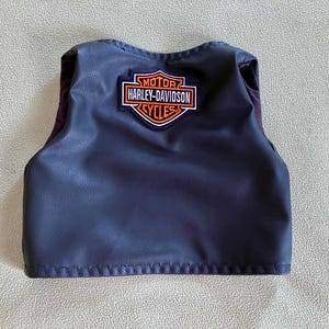 Puede incluir: Un peque&ntilde;o chaleco azul oscuro con el parche del logotipo de Harley-Davidson. El parche es rectangular con las palabras "MOTOR HARLEY-DAVIDSON CYCLES" en naranja y blanco. El chaleco tiene aspecto de cuero.