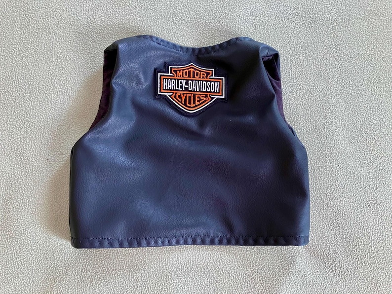 Puede incluir: Un peque&ntilde;o chaleco azul oscuro con el parche del logotipo de Harley-Davidson. El parche es rectangular con las palabras "MOTOR HARLEY-DAVIDSON CYCLES" en naranja y blanco. El chaleco tiene aspecto de cuero.