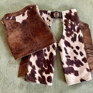 May include: A pair of miniature chaps, featuring a brown leather-like material and a cowhide pattern. The chaps have a belt with a buckle and fringe detailing. The overall design is reminiscent of Western wear.