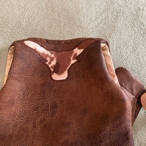 May include: A brown faux leather vest featuring a textured surface and a prominent longhorn steer head appliqu&eacute; in shades of brown and cream at the upper back. The garment includes a light beige lining and a silver-toned star-shaped metal accent.