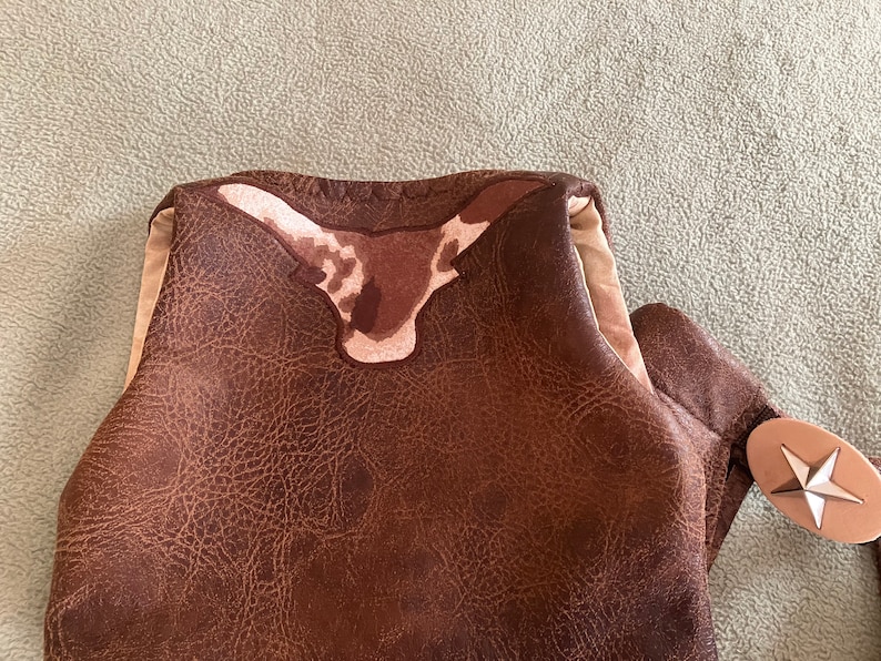 May include: A brown faux leather vest featuring a textured surface and a prominent longhorn steer head appliqu&eacute; in shades of brown and cream at the upper back. The garment includes a light beige lining and a silver-toned star-shaped metal accent.