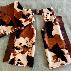 May include: A brown and white cow print chaps with fringe and a brown leather belt. The chaps are made of a soft, plush fabric.