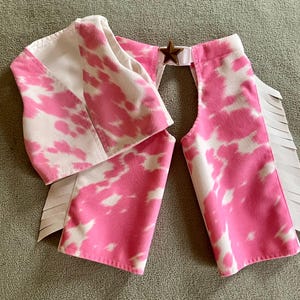 May include: A pink and white cow print vest with a star accent and white fringe. The vest is made of a soft material and is designed to be worn as a costume. The vest is a fun and playful accessory.