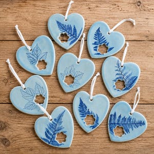 May include: Nine light blue heart-shaped ceramic ornaments, each featuring a dark blue botanical print of a fern or leaf and a small flower-shaped cutout. Each ornament includes a white string loop for hanging, arranged on a wooden surface.