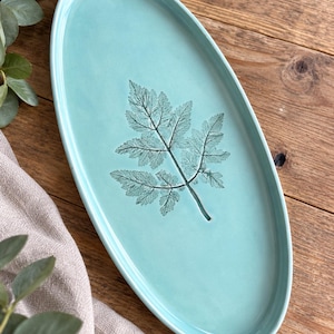 May include: An oval-shaped ceramic tray in a light turquoise colour featuring a detailed, dark green fern leaf imprint centred on its smooth surface. The tray has a raised rim and a glossy finish, showcasing a botanical design on the flat base.