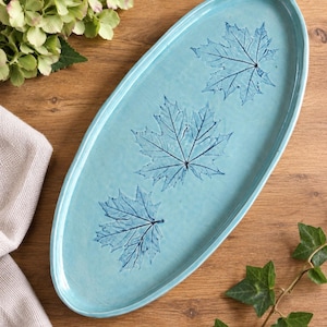 May include: An oval-shaped ceramic serving platter in a glossy light blue finish. The surface features three dark blue maple leaf imprints arranged in a diagonal line, showcasing detailed vein patterns pressed into the glazed ceramic material.
