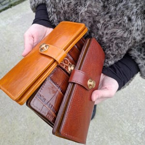 May include: Three rectangular wallets in varying shades of brown leather. One is a smooth tan, one features a dark brown crocodile-embossed texture, and the third is a textured reddish-brown. Each wallet has a flap closure secured with a brass button.
