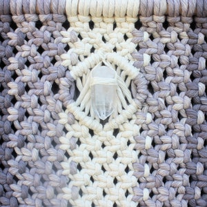 May include: A macrame wall hanging with a white crystal in the centre. The macrame is made with a gradient of white and grey yarn.