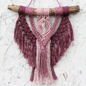 May include: A pink and white macrame wall hanging with a crystal in the center. The hanging is made with cotton cord and has a wooden dowel at the top.