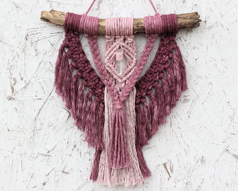 May include: A pink and white macrame wall hanging with a crystal in the center. The hanging is made with cotton cord and has a wooden dowel at the top.