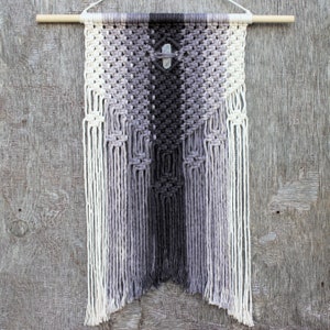 May include: A grey and white macrame wall hanging with a geometric pattern and a small white stone in the centre. The hanging is made with thick cotton cord and has long fringe on the bottom.