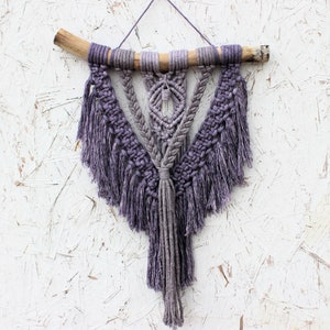 May include: A purple and gray macrame wall hanging with a wooden dowel at the top and fringe details.
