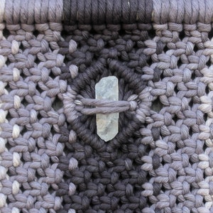 May include: A close-up of a woven wall hanging with a light blue crystal in the centre. The wall hanging is made of a gradient of grey yarn.