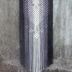 May include: A macrame wall hanging with a geometric pattern in shades of grey and white. A crystal is hanging in the centre of the design.