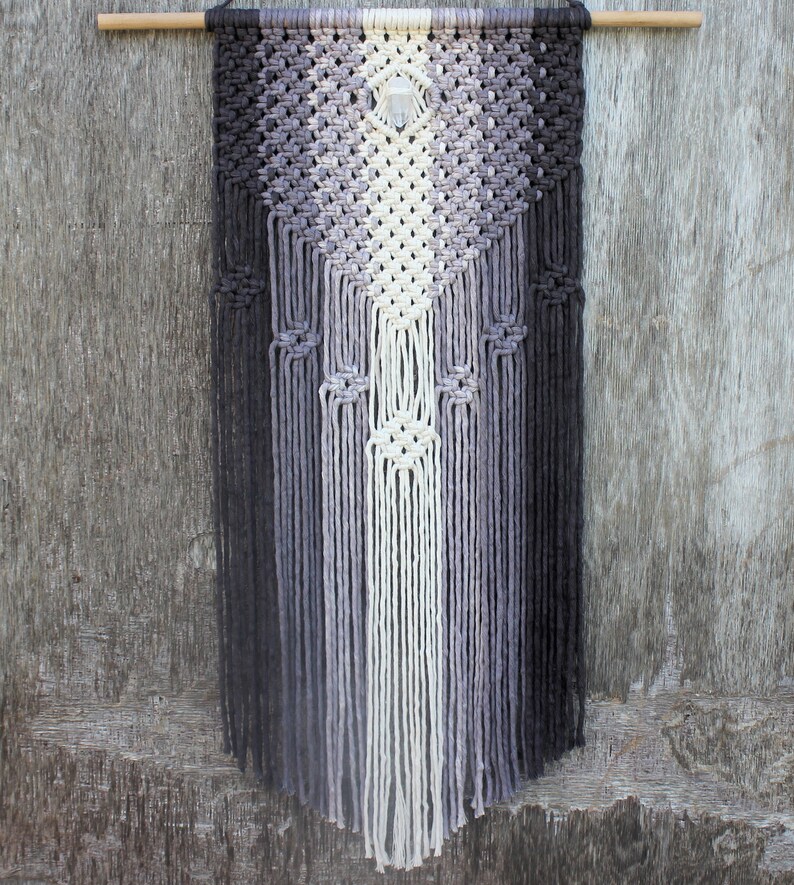May include: A macrame wall hanging with a geometric pattern in shades of grey and white. A crystal is hanging in the centre of the design.