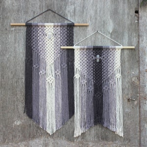 May include: Two macrame wall hangings with a geometric pattern in shades of grey and white. The larger hanging is slightly wider than the smaller hanging. Both hangings have a wooden dowel at the top and a crystal in the centre.